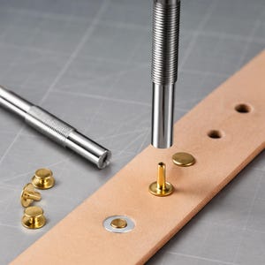 May include: Two silver-colored metal setting tools with knurled grips, several brass-colored metal rivets, and a light tan leather strip with pre-punched holes. One rivet is positioned upright on the leather, while another is set in a hole.