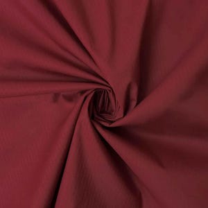 May include: A solid deep burgundy fabric with a smooth, matte texture. The material is gathered in the center, creating soft, radiating folds and shadows that highlight the consistent dark red color and the fine weave of the textile surface.