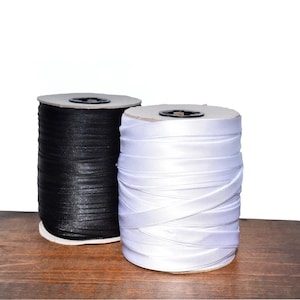 May include: Two cylindrical spools of ribbon, one in solid black and one in solid white. Each spool features a smooth, satin-like texture with the ribbon wound tightly around a central core, showing a consistent width and a reflective finish.