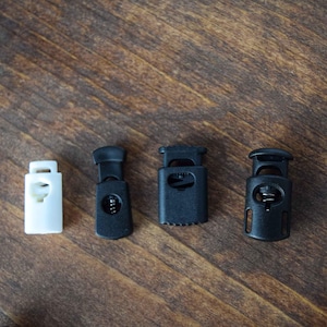 May include: A set of four cord locks, including one white plastic toggle and three black plastic toggles of varying shapes. Each toggle features a spring-loaded mechanism designed to secure drawstrings on clothing, bags, or outdoor gear.