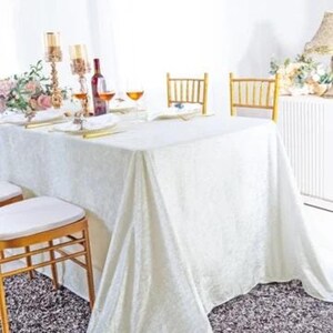 May include: A rectangular white tablecloth with a subtle damask pattern draped over a dining table. The table is set with gold-toned chairs, wine glasses, a wine bottle, and tall gold candle holders, creating a formal dining arrangement.