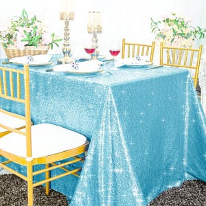 May include: A rectangular table covered with a bright light blue sequin tablecloth. The table is set with white plates, wine glasses, and tall metallic candle holders. Two gold-toned chairs with white cushions are positioned at the table.