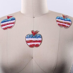 May include: Three apple-shaped patches featuring a sequined design with a blue top section containing silver stars, followed by horizontal stripes in white and red. Each patch has a small red sequined stem at the top, attached to a beige mannequin.