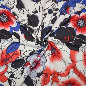 May include: A white fabric featuring a bold floral pattern with large, vibrant red and blue flowers. The design includes black outlines and solid black leaves, creating a high-contrast graphic print across the surface of the textile material.