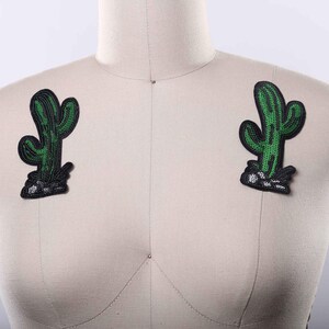 May include: Two matching cactus-shaped patches featuring vibrant green sequins arranged in a vertical ribbed pattern. Each patch has a black embroidered border and a base detailed with white and black sequins to represent rocky ground.