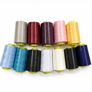 May include: A collection of twelve cylindrical spools of sewing thread arranged in two rows. The spools feature a variety of solid colors including light blue, teal, black, white, cream, dark gray, navy, gray, maroon, dark purple, pink, and yellow.