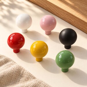 May include: A collection of six spherical cabinet knobs in solid colors including white, light pink, black, red, yellow, and green. Each knob features a smooth, glossy finish and a short cylindrical base for mounting to furniture or doors.