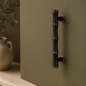 Puede incluir: Un tirador de armario vertical con acabado negro mate y un diseño texturizado inspirado en el bambú. El tirador cuenta con dos puntos de montaje circulares con detalles metálicos en tono bronce sobre una superficie verde oliva.