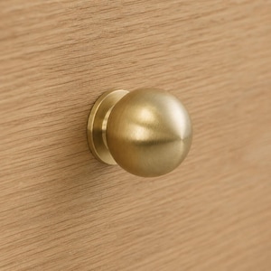 May include: A spherical cabinet knob with a brushed brass finish, featuring a smooth, rounded surface and a circular base plate. The knob is mounted onto a light-colored wooden surface with a visible horizontal grain pattern throughout the wood.