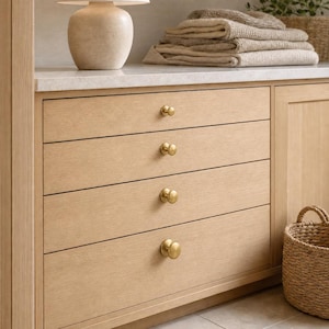 May include: A light wood cabinet featuring four horizontal drawers, each equipped with a round, brushed brass knob. The cabinet has a smooth, light-colored countertop surface, and a woven wicker basket sits on the floor to the right side.