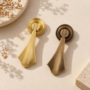 May include: Two metal drop pulls for furniture, one with a bright brushed gold finish and the other with a darker, antiqued brass finish. Each pull features a circular mounting plate and a flared, teardrop-shaped handle that hangs vertically.