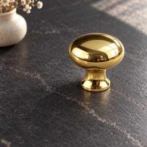 Könnte beinhalten: Ein polierter goldfarbener Möbelknopf aus Metall mit einem glatten, abgerundeten, pilzförmigen Kopf und einem konisch zulaufenden zylindrischen Sockel. Die Oberfläche des Knopfes ist hochglänzend und weist Lichtreflexe sowie ein Metallfinish auf.