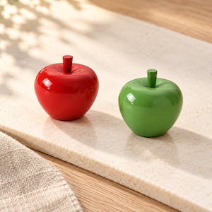 May include: Two smooth, glossy apple-shaped decorative objects. One apple is a vibrant, solid red color, and the other is a bright, solid green color. Both items feature a small, matching cylindrical stem at the top center.