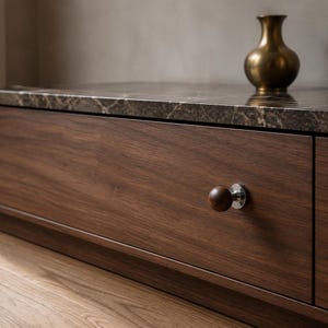 Puede incluir: Un frente de cajón de madera marrón oscuro con un pomo esférico de madera sobre una base de metal plateado pulido. El cajón está rematado con una superficie de mármol marrón oscuro con vetas blancas y un pequeño jarrón de color latón.