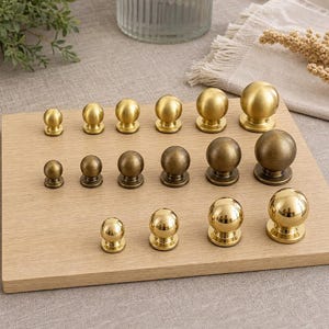 May include: A collection of sixteen spherical knobs arranged in three rows on a wooden board. The top row features six matte gold knobs, the middle row has six antique brass knobs, and the bottom row contains four polished gold knobs.