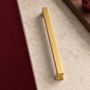 May include: A long, straight cabinet pull handle with a brushed gold finish. The hardware features a rectangular profile with slightly flared, concave ends, showcasing a smooth, metallic texture that reflects light along its central length.