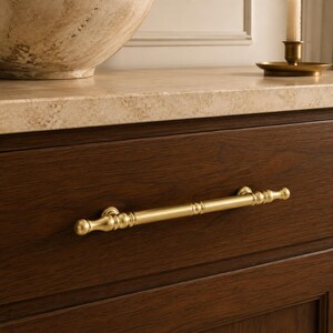 May include: A horizontal brass drawer pull with a smooth, cylindrical center and decorative rounded accents at each end and along the shaft. The hardware is mounted on a dark brown wooden drawer front with a visible grain, positioned below a stone countertop.