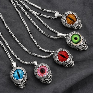 Peut inclure: Cinq pendentifs crânes en métal argenté sur des chaînes à maillons carrés assorties. Chaque crâne présente une surface texturée et vieillie avec un grand œil en verre réaliste au centre. Les yeux sont orange, verts, rouges, roses et bleus.