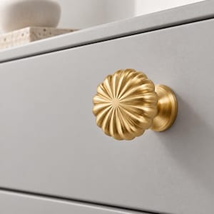 Puede incluir: Un pomo de mueble redondo de metal dorado con un diseño estriado en forma de pétalos que irradia desde el centro. El pomo presenta un acabado metálico cepillado y está montado en un frente de cajón gris claro liso con una costura horizontal.