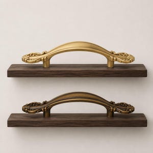 May include: Two arched metal drawer pulls with ornate, scroll-like detailing at each end. The top pull has a smooth, matte gold finish, while the bottom pull features a darker, antique bronze finish. Each pull is mounted on a dark wood block.