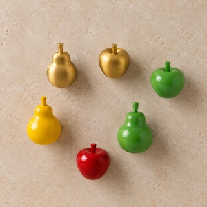 May include: A collection of six fruit-shaped knobs or decorative objects, including a gold pear, a gold apple, a bright green apple, a yellow pear, a red apple, and a green pear, all featuring smooth, solid-colored finishes and stems.