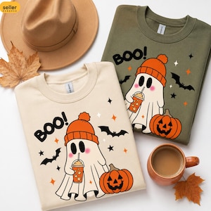 May include: A beige sweatshirt and an olive green sweatshirt, each featuring a graphic of a ghost wearing an orange beanie, holding an iced coffee, and standing next to a jack-o'-lantern with bats and the text BOO printed in black.