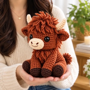 May include: A crochet stuffed animal shaped like a Highland cow with shaggy dark brown yarn, a light beige muzzle, two small cream-coloured horns, and large black plastic eyes. The toy features a textured, knotted yarn finish and dark brown hooves.