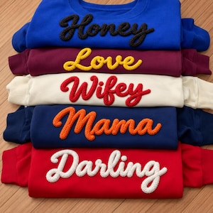 Könnte beinhalten: Ein Stapel aus fünf langärmeligen Sweatshirts mit Rundhalsausschnitt in Blau, Kastanienbraun, Creme, Marineblau und Rot. Jedes Sweatshirt hat ein strukturiertes, erhabenes Schriftwort auf der Brust: Honey in Schwarz, Love in Gelb, Wifey in Rot, Mama in Orange und Darling in Weiß.