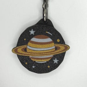 May include: A circular keychain featuring a glittery black background with a depiction of a planet with rings. The planet has horizontal bands of brown, white, and gold glitter. Small white stars and gold dots are scattered across the black space.