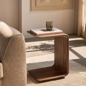 May include: A C-shaped side table crafted from smooth, medium-toned wood with rounded edges. A light-coloured book and a small, ribbed, translucent brown glass tumbler are placed on the flat top surface of the table.