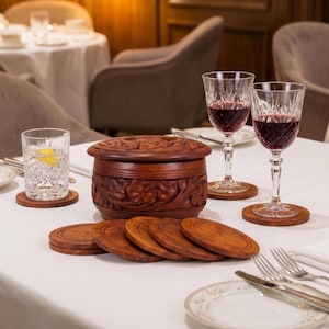 May include: A round, dark brown wooden container with a carved floral pattern on the lid and sides, accompanied by a set of matching circular wooden coasters. The set is arranged on a white tablecloth alongside crystal glassware filled with red wine.