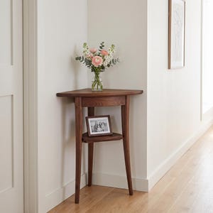 Puede incluir: Una mesa esquinera de madera oscura con una superficie triangular, un cajón y un estante inferior. Un florero de vidrio transparente con flores rosas y blancas se encuentra sobre la superficie, y un pequeño marco de fotos rectangular descansa en el estante.