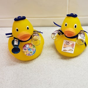 May include: Two yellow rubber duckies wearing blue yarmulkes and blue ribbons. One duck has a small bottle of wine and a small jar of gefilte fish. The other duck has a small bottle of wine and a package of matzah.