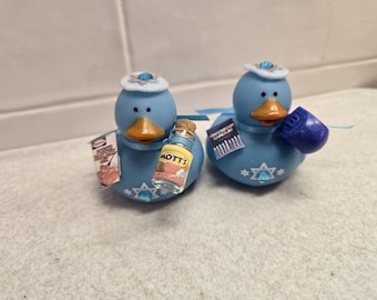 Könnte beinhalten: Zwei blaue dekorative Entenfiguren mit orangefarbenen Schnäbeln und weißen Hüten. Eine Ente hält eine kleine Flasche mit der Aufschrift "MOTTS" und ein kleines Schild. Die andere Ente hält ein kleines blaues Objekt und ein Schild mit der Aufschrift "CHANUKAH CANDLES".