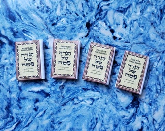 May include: Four miniature Passover Haggadah books with Hebrew text on the cover. The books are white with brown trim and are arranged in a row on a blue and white marble surface.