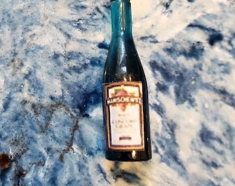 May include: A miniature glass bottle with a red cap and a white label. The label has black text that reads "KING'S OWN" and "GRAPE" in a cursive font. The bottle is filled with a dark liquid.