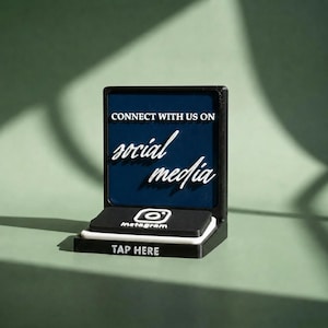 Puede incluir: Un soporte de exhibición cuadrado negro con un panel azul oscuro con texto blanco que dice CONNECT WITH US ON social media. Debajo, una plataforma negra muestra un logotipo de Instagram blanco y el texto TAP HERE en letras blancas.
