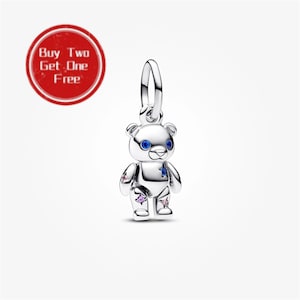May include: A silver teddy bear charm featuring blue gemstone eyes and small star-shaped accents in blue and pink on its body. The charm includes a circular bail at the top for attachment. A red circular badge with the text Buy Two Get One Free is visible.