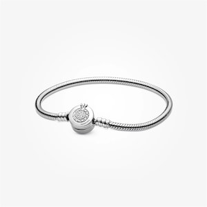 May include: A silver snake chain bracelet featuring a round barrel clasp. The clasp is decorated with a circular pavé setting of clear stones and the engraved text PANDORA. The bracelet has a smooth, flexible metallic finish throughout.
