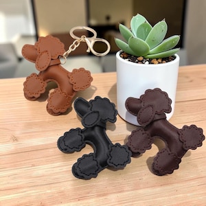 May include: Three poodle-shaped keychains in tan, black, and dark brown leather-like material. Each keychain features scalloped edges on the ears, tail, and feet, with a gold-toned metal split ring and lobster clasp attached to the tan poodle.