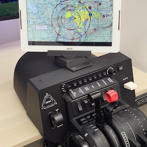 Può includere: Una console di controllo per simulatore di volo nera con leva del carrello, manette dell'acceleratore e vari interruttori etichettati per avionica, luci di atterraggio e flap. Un tablet Acer mostra una carta di navigazione aeronautica.