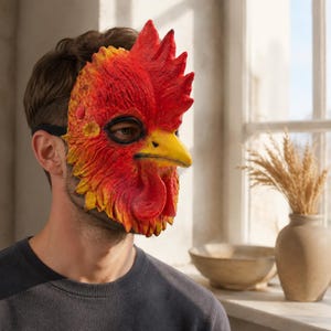 Puede incluir: Una máscara de gallo moldeada con una textura de plumas de colores rojo y amarillo vibrantes, una cresta roja prominente y un pico amarillo. La máscara cubre la parte frontal del rostro y se asegura con una banda elástica negra, mostrando detalles.