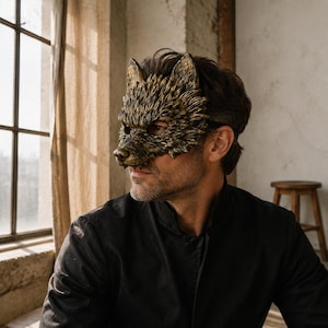 May include: A sculpted wolf mask featuring a textured, metallic gold and black finish with detailed fur-like carvings. The mask covers the upper portion of the face, including the eyes and nose, and is secured with a thin black strap around the head.