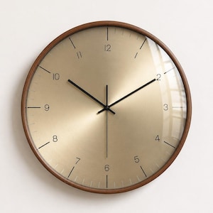 May include: A round wall clock with a dark brown wooden frame and a metallic gold face. The clock features thin black hands and black numerals for 2, 3, 4, 5, 6, 7, 8, 9, 10, and 12, with simple black tick marks for the remaining hour positions.