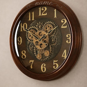 May include: A round wall clock with a dark brown wooden frame and a glass cover. The clock face features gold-coloured numbers from 1 to 12, exposed mechanical gears in the centre, and the word name printed in cursive at the top centre position.