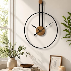 Op de afbeelding: Een wandklok met een groot zwart cirkelvormig metalen frame en een verticale steunbalk die uitloopt in een klein houten accent aan de bovenkant. Het midden heeft een ronde houten wijzerplaat met zwarte metalen wijzers en een ster.