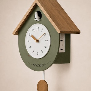 May include: A house-shaped wall clock with a matte olive green body and a natural wood roof. The white circular clock face features black numerals and gold hands. A small white bird sits above the dial, and a wooden pendulum hangs below.