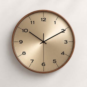 May include: A round wall clock with a dark brown wooden frame and a metallic gold face. The clock features black Arabic numerals from 1 to 12, thin black hour and minute hands, and a thin black second hand centred on the dial.