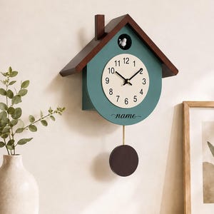 May include: A house-shaped wall clock with a teal body and a dark brown roof. The clock face is cream-coloured with black numerals and hands. A small white bird figure sits in an opening above the dial. The word name is printed below the dial.