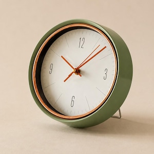 May include: A round analog table clock with a sage green outer casing and a metallic copper-toned inner rim. The clock face is white with black numerals at 3, 6, 9, and 12, featuring thin black tick marks and three copper-colored hands.